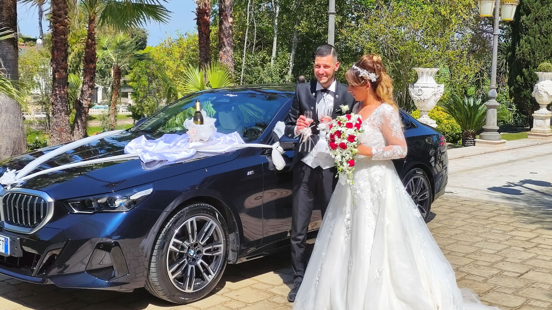 Sposi felici brindano con champagne accanto a una lussuosa auto addobbata della Roman Limousine Service, con una bottiglia di champagne elegantemente posizionata sul cofano.