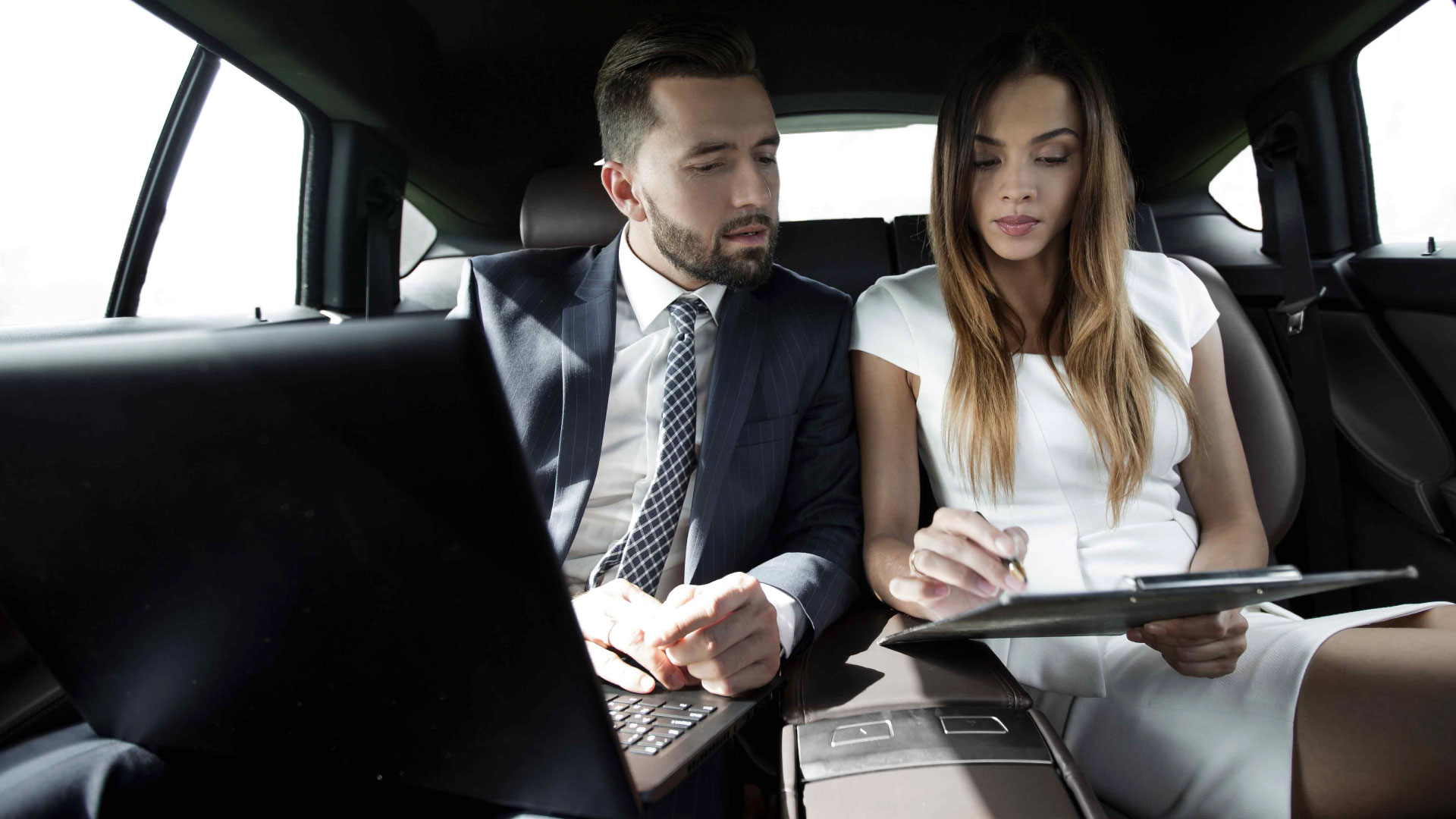 Due professionisti, un uomo e una donna, impegnati in una discussione di lavoro con laptop e blocco note, comodamente seduti all’interno di un’auto di lusso fornita da Roman Limousine Service.